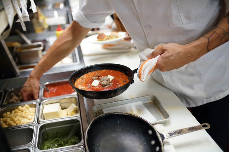 chef holding a skillet with red sauce and meatballs in it adding more ingredients